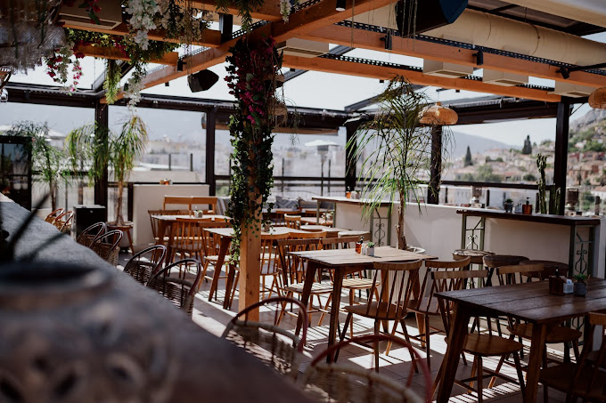 Rooftop bar and restaurant with wooden tables and chairs, decorated with hanging plants and flowers. The open-air setting features a view of the city in the background, creating a relaxed and inviting atmosphere with natural light and greenery enhancing the ambiance