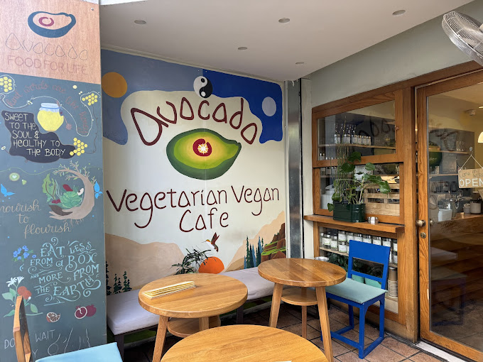 Entrance of 'Avocado Vegetarian Vegan Cafe,' featuring colorful murals and cozy wooden tables with chairs. The mural includes an avocado illustration and cheerful messages promoting plant-based eating, with potted plants adding to the welcoming, natural atmosphere