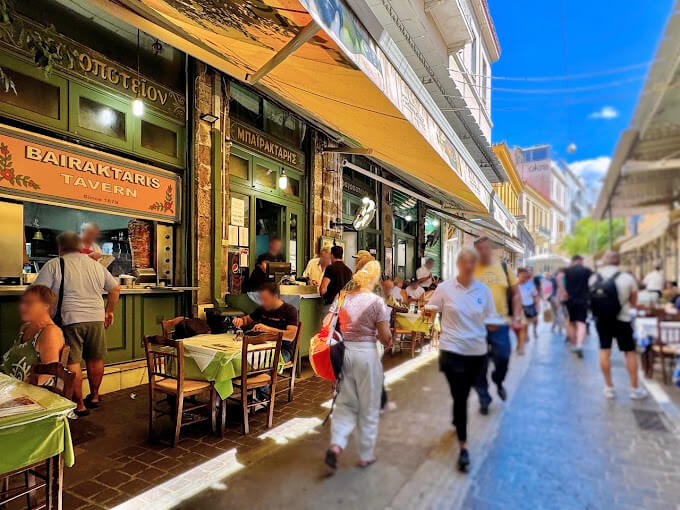 Street view of 'Bairaktaris Tavern' in Monastiraki, Athens, with people walking by and diners seated at tables covered in green tablecloths. The vibrant scene captures the bustling energy of a traditional Greek taverna, with an open kitchen and a large awning shading the outdoor seating area