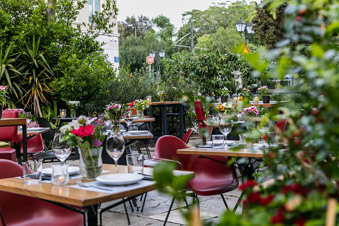 Charming outdoor dining area with wooden tables, red chairs, and lush greenery surrounding the space. Tables are set with glassware, plates, and small vases of colorful flowers, creating a vibrant and inviting garden atmosphere for dining al fresco