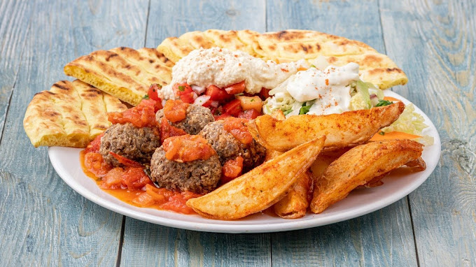 Plate of Greek-style food featuring grilled pita bread, meatballs topped with tomato sauce, seasoned potato wedges, and a side salad with a dollop of tzatziki. The dish is vibrant and hearty, served on a rustic blue wooden table