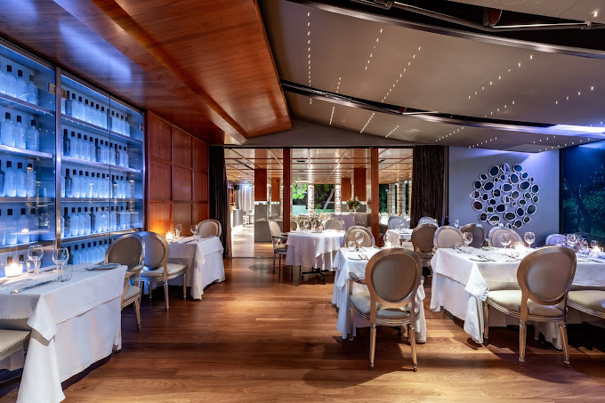 Elegant restaurant interior featuring round tables covered with white tablecloths, set with fine dining place settings. The space has soft ambient lighting, a contemporary design with wooden accents, and modern decor, including illuminated shelving with bottles and a decorative wall installation, creating a sophisticated and upscale atmosphere