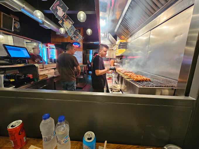 View of a souvlaki shop kitchen with a cook grilling skewers over an open flame. The scene shows the bustling atmosphere, with skewers lined up on the grill, and other staff members working in the background. Bottled water and soda cans are visible on the counter in the foreground