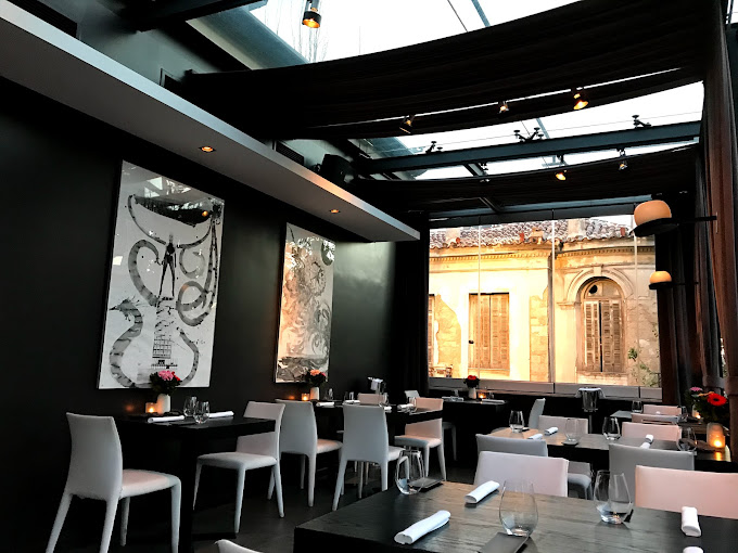 Chic, modern dining room with dark walls and white minimalist furniture, featuring contemporary artwork and a partially open ceiling with draped fabric. The large window offers a view of an adjacent historic building, blending a sophisticated interior ambiance with urban charm