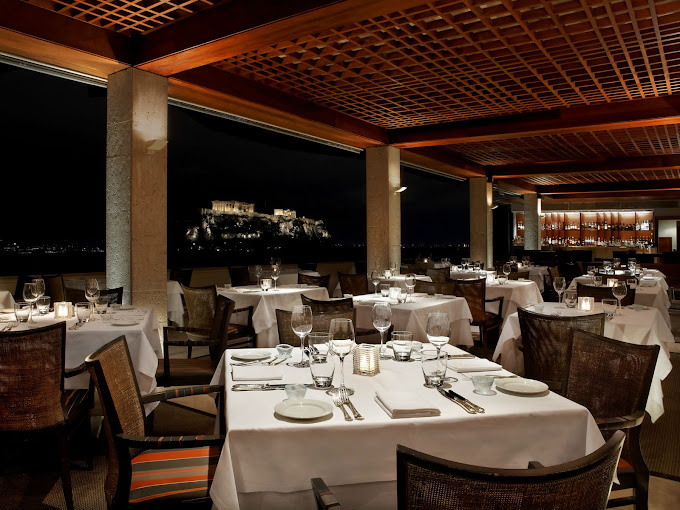 Elegant rooftop restaurant dining area with neatly set tables, overlooking the Acropolis at night. The space features soft ambient lighting, white tablecloths, glassware, and a warm wooden ceiling, creating a sophisticated and romantic atmosphere with a stunning view of Athens' iconic landmark