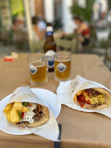 Two gyro wraps filled with meat, French fries, tomatoes, and sauce, served on a table with two glasses of beer and a beer bottle in the background. The scene is set outdoors, with a relaxed atmosphere suggesting casual street dining