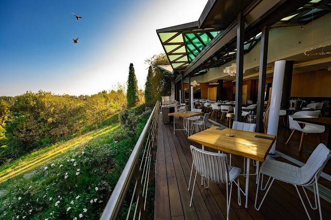 Outdoor terrace of a restaurant overlooking a lush green landscape, with wooden tables and modern wire chairs arranged along a deck. The open-air setting is complemented by an expansive view of nature, under a partially covered roof with glass panels, creating a tranquil and scenic dining experience