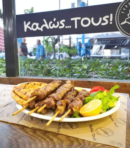 Plate of skewered souvlaki served with grilled pita bread, a lemon wedge, and fresh greens, placed on a branded placemat. In the background, a sign reads 'Καλώς...ΤΟΥΣ!' (Kalos... Tous!), adding a welcoming touch, with a window view of the street outside