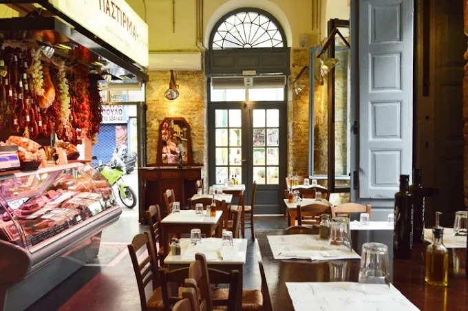 Interior of a traditional delicatessen-style restaurant featuring wooden tables set with simple glassware and condiments. The space has a charming rustic feel, with hanging cured meats displayed near a glass deli counter, exposed stone walls, and tall windows that let in natural light