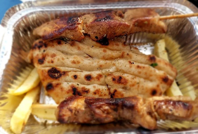 Takeout container with grilled skewered meat (souvlaki) placed on top of a piece of pita bread, with French fries underneath. The dish is served in a foil tray, with the pita and meat showing grill marks, giving a classic Greek street food appearance