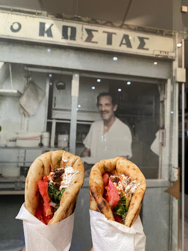 Two gyros wrapped in pita bread filled with tomatoes, herbs, and sauce, held up in front of an old photograph of a man smiling, with a sign above reading 'Ο Κώστας' (O Kostas). The background suggests a nostalgic connection to a well-known Greek eatery