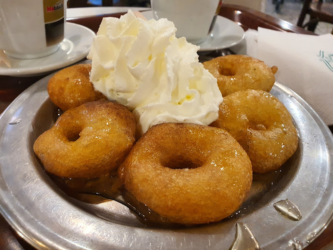 Plate of loukoumades, traditional Greek doughnuts, drizzled with honey and topped with a generous dollop of whipped cream. The golden-brown pastries are glistening with syrup, served alongside cups of coffee in the background, creating a classic Greek dessert setting