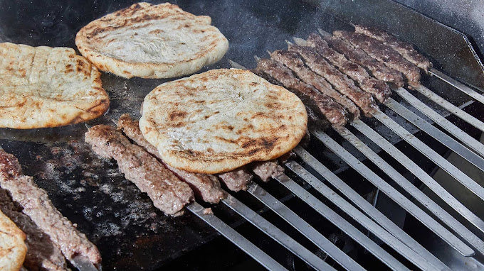 Close-up of kebabs grilling on skewers alongside pieces of pita bread on a hot grill. The skewers are lined up, and the pita is toasting, with steam rising, showcasing the preparation of traditional Greek street food