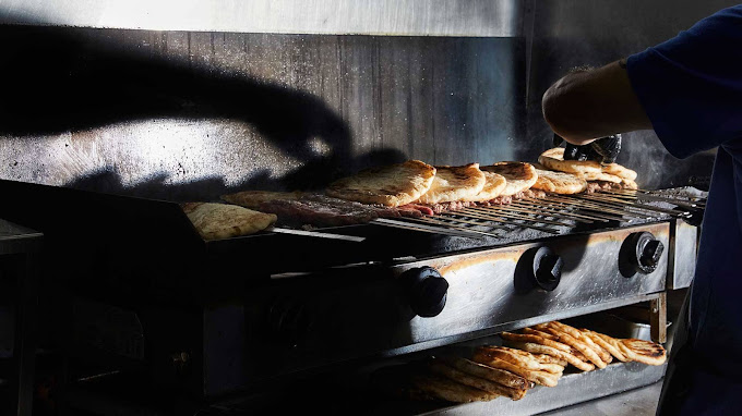 Dimly lit scene of pita bread grilling on a charcoal grill, with a cook's hand reaching to adjust the bread. The grill is filled with rows of pita being toasted, with some stacked underneath, highlighting the traditional preparation of Greek street food