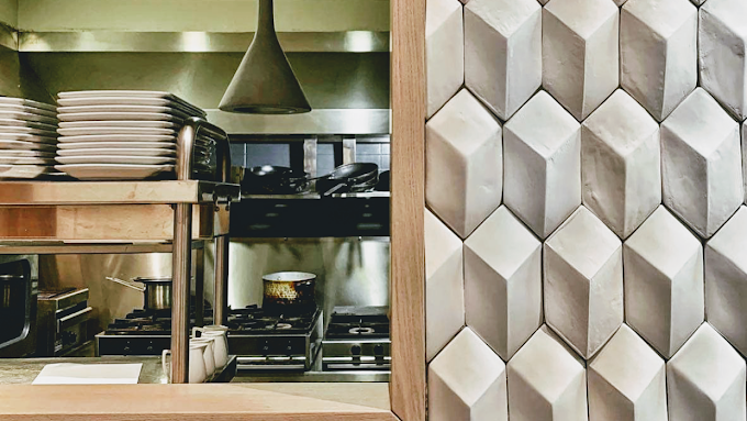 View of a restaurant kitchen through a service window, with stacked plates, cooking utensils, and a stove in the background. The right side of the image features textured, geometric wall tiles, adding a stylish design element to the scene