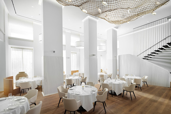 Bright, elegant restaurant interior with high ceilings and round tables covered in white tablecloths. The room features minimalist cream-colored chairs, large pillars, and an artistic ceiling installation resembling a fishing net. A staircase with sleek black railings adds to the contemporary, airy atmosphere