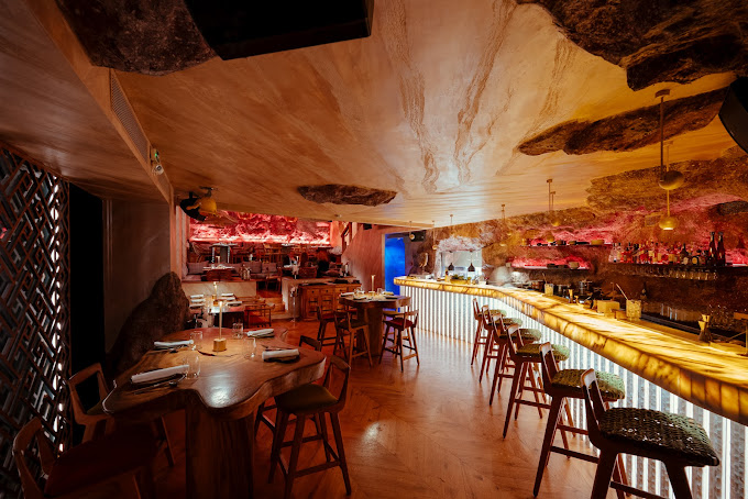 Stylish, dimly lit restaurant interior featuring an illuminated bar counter with high stools, natural wood tables, and a ceiling designed with rugged stone accents. The warm ambient lighting creates an intimate and exotic atmosphere, with red hues adding a dramatic touch to the unique decor