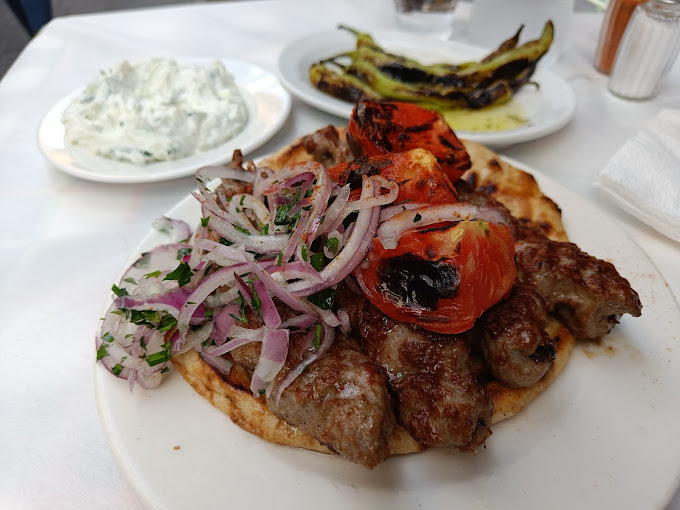 Plate of grilled kebabs served on pita bread, topped with sliced red onions and roasted tomatoes. Accompanying side dishes include a plate of charred green peppers and a serving of tzatziki, all arranged on a white tablecloth, giving a classic Greek meal presentation