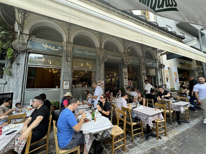 Outdoor seating area of 'O Thanasis' restaurant in Monastiraki, Athens, with customers enjoying their meals at tables covered in checkered tablecloths. The bustling atmosphere features people dining under a large awning, with the classic facade of the restaurant visible in the background