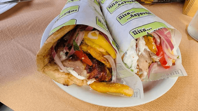 Close-up of two gyro wraps on a plate, filled with grilled meat, French fries, tomatoes, onions, and tzatziki. The wraps are partially covered with branded paper, showcasing a hearty and colorful Greek street food meal