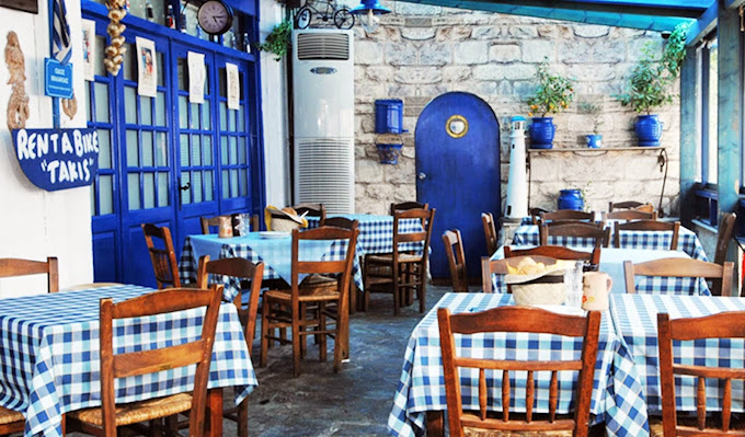 Traditional seafood Greek taverna with blue and white checkered tablecloths, wooden chairs, and vibrant blue decor. The space has a rustic charm, featuring stone walls, potted plants, and a cozy atmosphere, with a sign reading 'Rent a Bike Takis' on the left