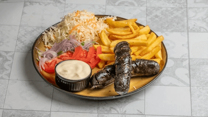 Plate of traditional Greek food featuring sausages, French fries, shredded cabbage and carrot salad, sliced tomatoes, and red onions, served with a small bowl of creamy dipping sauce. The dish is arranged on a decorative plate, showcasing a variety of textures and flavors