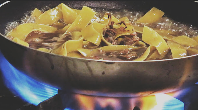 Close-up of wide ribbon pasta cooking in a frying pan over an open flame. The dish features a rich broth with pieces of meat, visibly bubbling and sizzling, showcasing the process of cooking pasta in a flavorful sauce