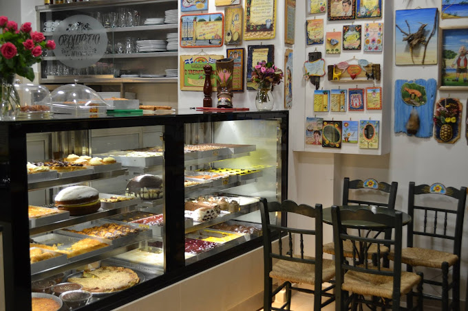 Cozy dessert shop interior featuring a glass display case filled with a variety of pastries and cakes. The walls are adorned with colorful vintage posters and decorations, and there are several wooden chairs with woven seats arranged nearby, creating a warm, nostalgic ambiance