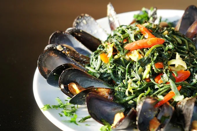 Close-up of a seafood dish featuring cooked mussels arranged around a bed of sautéed greens, garnished with slices of red and yellow peppers. The dish appears fresh and vibrant, showcasing a mix of shellfish and vegetables