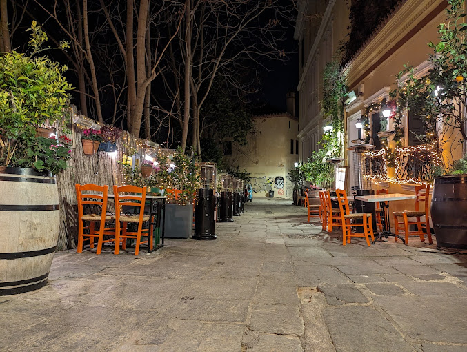 Outdoor dining area of a cozy taverna at night, featuring orange wooden chairs and tables along a stone-paved alley. The space is adorned with potted plants, string lights, and greenery, creating an inviting atmosphere, with leafless trees and soft ambient lighting enhancing the charming setting