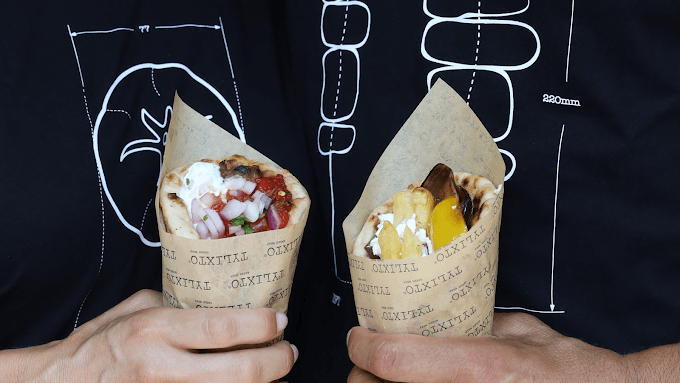 Close-up of two hands holding Greek wraps in paper sleeves. The wraps are filled with ingredients like tomatoes, onions, tzatziki, fries, and roasted vegetables, against a background featuring a diagram-style graphic on a black shirt