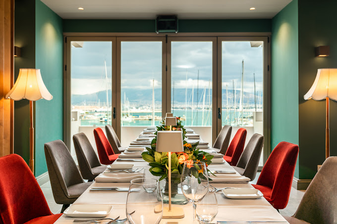 Elegant dining room with a long table set for a meal, featuring neatly arranged plates, glassware, and floral centerpieces. The room has a view of a marina through large windows, with sailboats visible in the distance, complemented by soft lighting from standing lamps and a cozy, upscale atmosphere