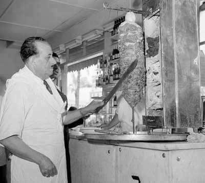 Black and white photo of a chef in a traditional kitchen slicing meat from a vertical rotisserie, preparing gyro. The setting is classic, with the chef wearing an apron and using a large knife, capturing a historical moment in Greek street food preparation