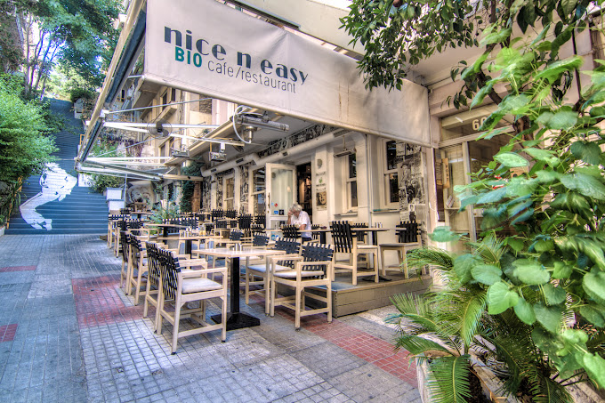 Outdoor seating area of 'nice n easy BIO cafe/restaurant,' featuring wooden tables and chairs arranged on a sidewalk. The exterior is adorned with greenery, and a large awning displays the restaurant's name. The atmosphere is relaxed and inviting, with a mural of a person visible on a nearby wall